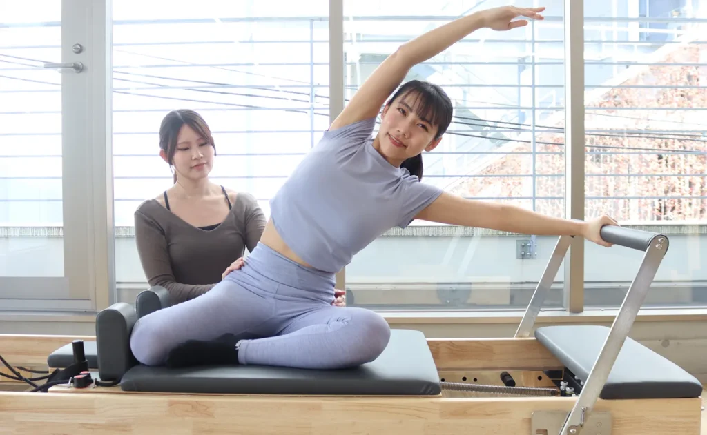 韓国式マシンピラティスでタワーリフォーマーを使用したストレッチを行う女性と、それをサポートするインストラクター