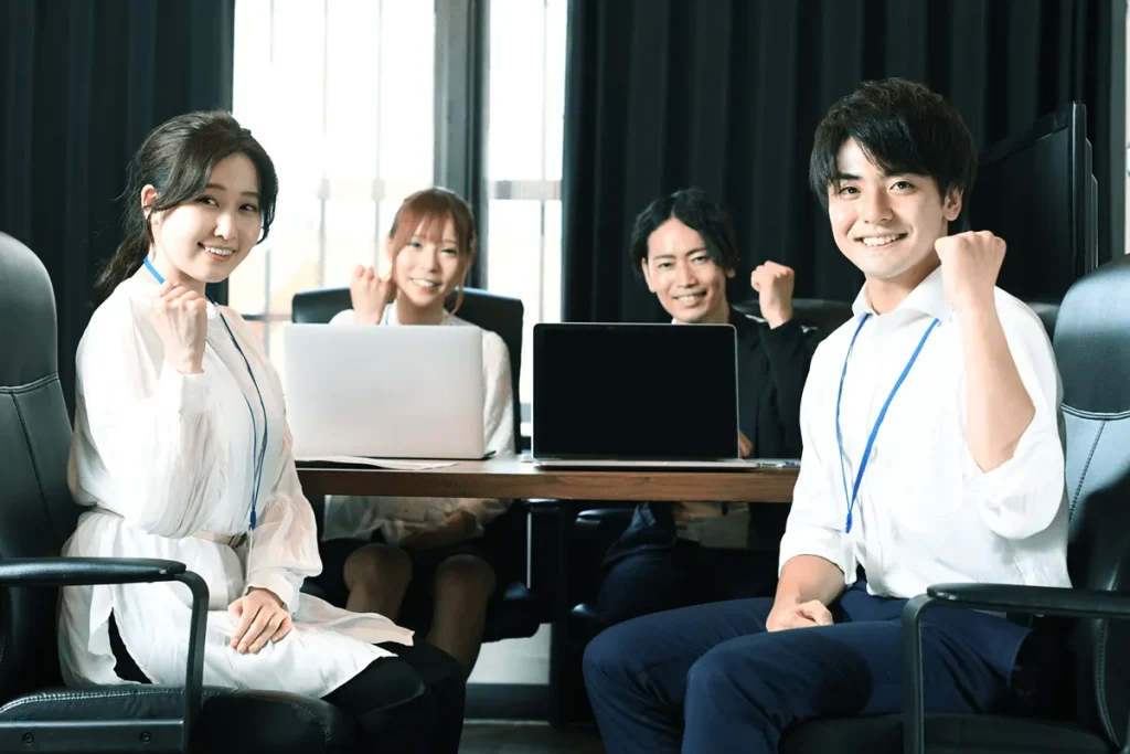 若手ビジネスチームが会議室でガッツポーズをしている様子。ポジティブな雰囲気の社内ミーティング